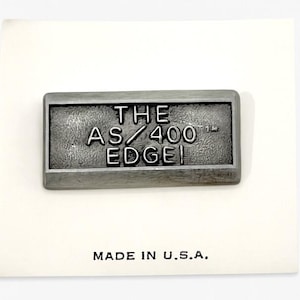 May include: A rectangular, silver-colored metal pin with the text "THE AS/400 EDGE!" in raised lettering. The pin is set against a white background with the words "MADE IN U.S.A." printed below.