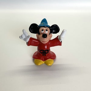 May include: A small plastic figurine of Mickey Mouse dressed as a sorcerer. He is wearing a red robe with a black sash, a blue pointed hat, and white gloves. He has a big smile and is holding his hands up in the air.