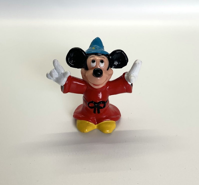 May include: A small plastic figurine of Mickey Mouse dressed as a sorcerer. He is wearing a red robe with a black sash, a blue pointed hat, and white gloves. He has a big smile and is holding his hands up in the air.