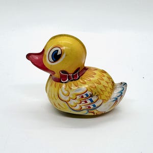 May include: A yellow wind-up toy duck with red, blue, and white details. The duck has a red beak and a bow tie around its neck.
