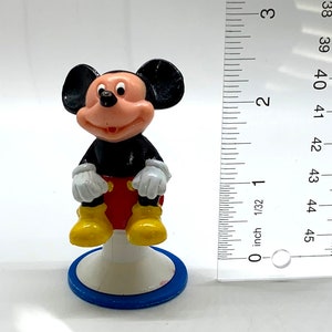 May include: A small plastic figurine of Mickey Mouse sitting on a blue and white base. Mickey is wearing his signature red shorts, yellow shoes, and white gloves.