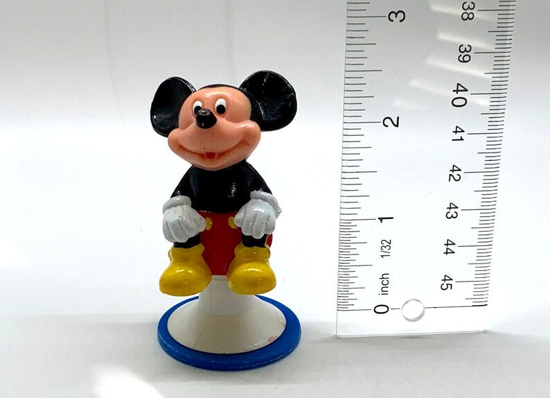 May include: A small plastic figurine of Mickey Mouse sitting on a blue and white base. Mickey is wearing his signature red shorts, yellow shoes, and white gloves.