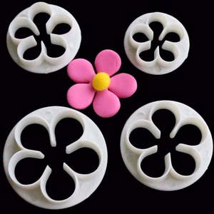 Op de afbeelding: Vier witte plastic bloemvormige koekjesuitstekers, twee grote en twee kleine, met een roze fondantbloem in het midden.