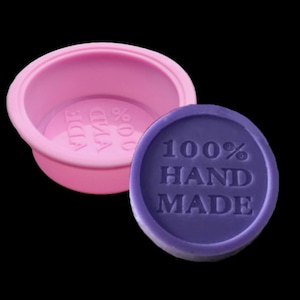 Puede incluir: Una pastilla de jab&oacute;n redonda de color morado con el texto "100% HAND MADE" en letras blancas. El jab&oacute;n est&aacute; situado junto a un molde de silicona rosa.