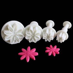 May include: Set of four white plastic flower-shaped cookie cutters with a pink fondant flower on a black background.