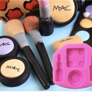 May include: Pink silicone mold for making makeup-themed cake decorations. The mold has four sections: a lipstick, a compact, and two bottles of nail polish. The mold is on a white surface.