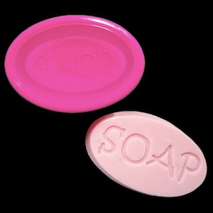 May include: A pink oval soap mold with the word "SOAP" embossed on it. Next to it is a pink oval bar of soap with the word "SOAP" embossed on it.