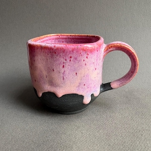 May include: A ceramic mug with a rounded square rim and a single handle. The upper portion features a glossy, mottled pink and magenta glaze that drips down over the lower section, which is finished in a matte, textured charcoal black glaze.