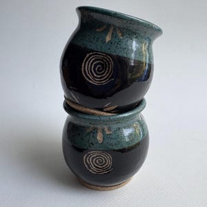May include: Two small, stacked ceramic pots. The pots are dark blue with a teal band near the top, decorated with a spiral design and leaf-like accents. The pots have a rustic, handmade appearance.