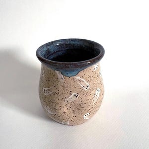 May include: A ceramic mug with a speckled beige body featuring a pattern of small white ghosts with black eyes. The rim of the mug is finished with a dark blue glaze that drips slightly down the exterior, contrasting with the speckled surface.
