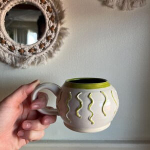 May include: Cream-colored ceramic mug with a rounded shape and a white handle. The mug features green wavy lines and a green interior rim. The mug is held in a hand.