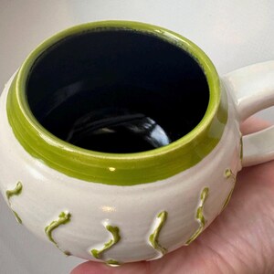 May include: A handmade ceramic mug with a white exterior, a vibrant green rim, and a dark blue interior. The mug features raised, organic green designs and a curved handle. This unique mug is perfect for coffee or tea.