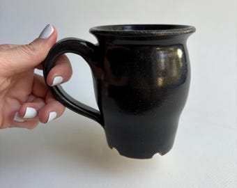 May include: A black ceramic mug with a rounded body and a sturdy handle. The mug has a slightly flared rim and a textured surface. The dark color and simple design give it a rustic, handmade feel. The mug is held by a hand with white nail polish.