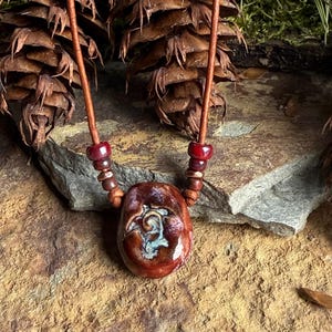 Può includere: Una collana in corda di cuoio marrone con perline rosse e marroni e un ciondolo in ceramica con un design di tartaruga bianca.