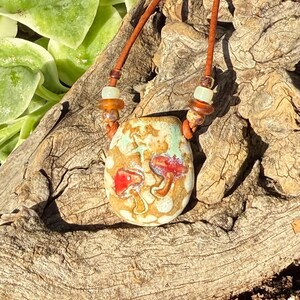 May include: A ceramic pendant necklace featuring a tan and light green speckled glaze with two hand-painted red mushrooms. The pendant hangs from a brown leather cord accented with small, translucent amber and pale green glass beads.