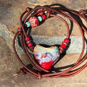 May include: A brown leather cord necklace featuring a central heart-shaped ceramic pendant with a marbled red, white, and turquoise glaze. The cord is accented with smaller red, turquoise, and copper-toned beads arranged symmetrically on both sides.