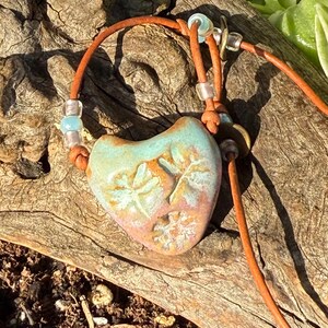 Puede incluir: Un colgante de cerámica en forma de corazón con esmalte turquesa y rosa, decorado con patrones en relieve de libélulas y copos de nieve. El colgante cuelga de un cordón de cuero marrón con cuentas de vidrio transparentes y azul claro.