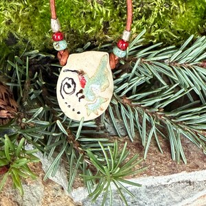Pode incluir: Um colar com um pingente de cerâmica com um design de beija-flor. O pingente é branco com detalhes em verde e vermelho. O colar é feito com cordão de couro marrom e contas vermelhas e azuis.