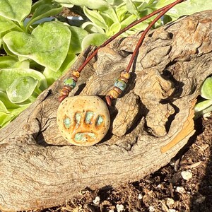 Puede incluir: Un collar con colgante de cerámica que presenta una piedra redonda de color beige moteado con un diseño de huella de pata esmaltado en turquesa. El colgante cuelga de un cordón de cuero marrón con pequeñas cuentas de cerámica a juego.