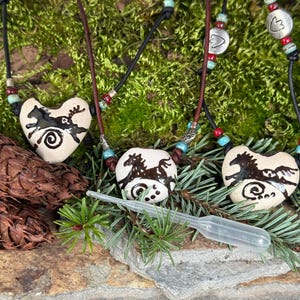 May include: Three ceramic heart-shaped pendants with black horse designs on a white background. Each pendant is on a black leather cord with turquoise and red beads. The pendants are displayed on a bed of moss.