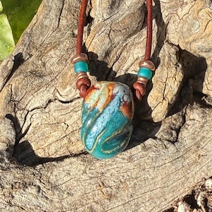 Può includere: Una collana con un ciondolo centrale in ceramica caratterizzato da uno smalto lucido con motivi vorticosi nei colori turchese, marrone e bianco. Il ciondolo è sospeso su un cordino in pelle marrone scuro, con piccole perline turchesi e color rame.