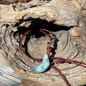 May include: A necklace featuring a brown leather cord with a curved, turquoise-colored ceramic pendant. The cord is accented with small, metallic-toned beads and a knot detail, creating a simple design that hangs in a circular loop.