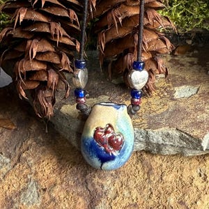 Może przedstawiać: Niebieski i brązowy ceramiczny wisiorek z czerwonym sercem, zawieszony na czarnym sznurku z srebrnymi i niebieskimi koralikami.