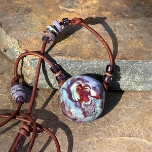 Pode incluir: Um colar com um pendente de cerâmica redondo e multicolorido, apresentando espirais de esmalte azul, roxo e castanho-avermelhado. O pendente está enfiado num cordão de couro castanho, acentuado com pequenas contas roxas e prateadas.