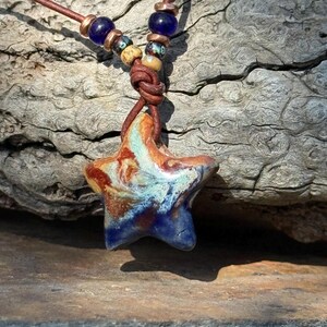 May include: A star-shaped ceramic pendant with a marbled glaze of blue, brown, and cream colors. The pendant hangs from a brown leather cord, which is accented with two dark blue glass beads and two smaller, multi-colored patterned beads.