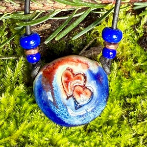 Może przedstawiać: Niebieski i czerwony ceramiczny wisiorek z wirującym wzorem zawieszony na czarnym skórzanym sznurku z niebieskimi i miedzianymi koralikami.