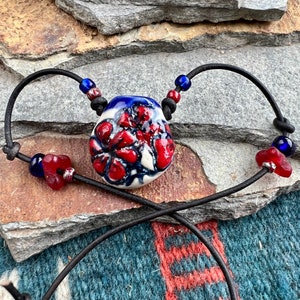 Puede incluir: Un colgante de cerámica rojo, blanco y azul con un diseño floral cuelga de un cordón de cuero negro con cuentas azules y rojas.
