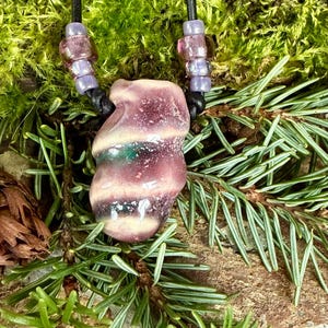 Może przedstawiać: Fioletowo-biały szklany wisiorek w kształcie spirali na czarnym skórzanym sznurku z fioletowymi szklanymi koralikami. Wisiorek jest unikatową i ręcznie robioną biżuterią.