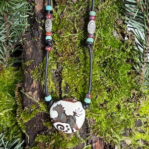 Puede incluir: Un collar artesanal con un colgante redondo blanco roto con un diseño de caballo marrón y verde. El colgante cuelga de un cordón negro, adornado con cuentas turquesas, rojas y plateadas. El collar se exhibe sobre una superficie de madera cubierta de musgo.