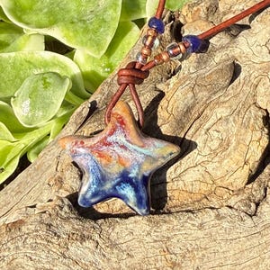 May include: A star-shaped pendant with a glossy, multi-colored glaze featuring swirls of deep blue, white, orange, and brown. The pendant hangs from a brown leather cord accented with small, translucent, and dark blue glass beads.