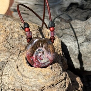 May include: A ceramic pendant featuring a spiral design in shades of red, brown, and grey, suspended on a dark brown leather cord. The cord is accented with small black, red, and translucent square beads positioned above the pendant.