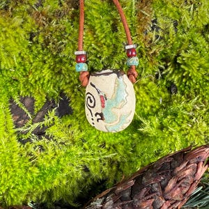 Puede incluir: Un colgante de cerámica con fondo blanco y diseños pintados en azul y rojo cuelga de un cordón de cuero marrón con cuentas rojas y azules. El colgante está sobre un lecho de musgo verde.