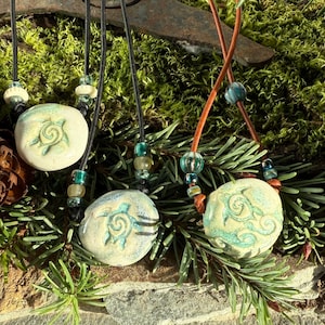 May include: Three handmade necklaces with round, off-white pendants featuring a teal spiral design. The pendants are strung on black and brown cords with colorful beads. The necklaces are displayed on a bed of greenery.
