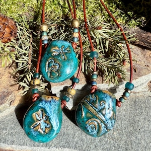 May include: Three turquoise blue ceramic pendants with gold accents and dragonfly designs hang from brown leather cords with turquoise and gold beads.