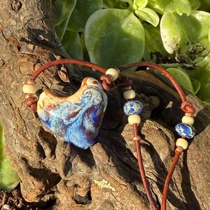 Puede incluir: Un collar con un colgante de cerámica en forma de corazón con un esmalte jaspeado en azul, blanco y marrón. El colgante está ensartado en un cordón de cuero marrón con pequeñas cuentas color crema y dos cuentas redondas de piedra azul y blanca.