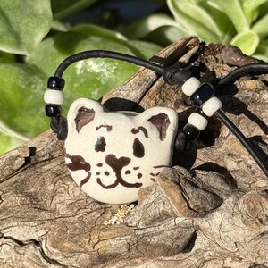 Pode incluir: Um pingente de cerâmica em formato de rosto de gato com base na cor creme e detalhes pintados em marrom escuro, incluindo olhos, nariz e bigodes. O pingente está preso a um cordão preto com contas cilíndricas alternadas em preto e branco.