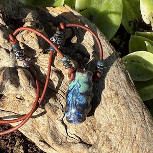 May include: A necklace featuring a glossy, organic-shaped ceramic pendant with a blue and green marbled glaze. The pendant hangs from a brown leather cord accented with small, round, black-and-white speckled beads and dark metallic-toned spacers.