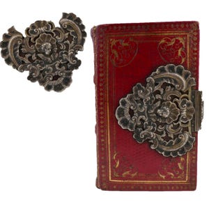 May include: A red leather-bound book with ornate gold detailing and two silver filigree clasps. The clasps are in the shape of a heart with floral designs.