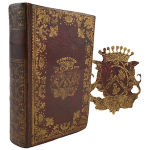 Può includere: Un libro rilegato in pelle marrone con decorazioni in foglia d'oro e un motivo floreale. Il libro ha uno stemma in foglia d'oro sulla copertina.