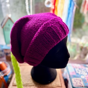 May include: A magenta knitted beanie hat on a black mannequin head. The hat features a ribbed brim and a relaxed fit. The background includes a window and decorative items.