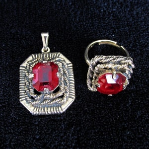 May include: A silver pendant and ring set featuring a red gemstone. The pendant has a square-shaped gemstone surrounded by a decorative silver frame. The ring has a similar design with a smaller gemstone.