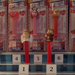 May include: A display of ten Valentine's Day themed Pez dispensers. The dispensers are arranged in two rows of five. The dispensers feature a variety of characters, including a heart, a ladybug, a bear, an angel, a monkey, and a cat. The dispensers are all red, pink, purple, or blue. The dispensers are labeled "To: My Valentine xoxo".