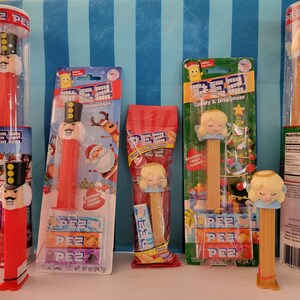 May include: Five Christmas-themed Pez dispensers, including a Nutcracker, an angel, and a Santa Claus. Each dispenser is packaged in a clear plastic tube or a colorful cardboard wrapper. The dispensers are all in red, white, and gold colors. The packaging includes the Pez logo and the words "Candy & Dispenser" and "Seasonal Candy Flavor".