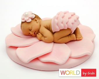 Può includere: Un topper per torta di pasta di zucchero rosa a forma di bambino che dorme su un fiore rosa. Il bambino indossa un pannolino rosa con volant e una fascia per capelli rosa con una perla.