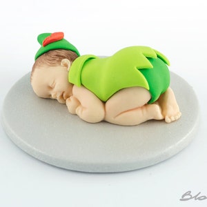 May include: A sleeping baby figurine made of fondant, wearing a green and red Peter Pan costume. The figurine is lying on a gray round base.