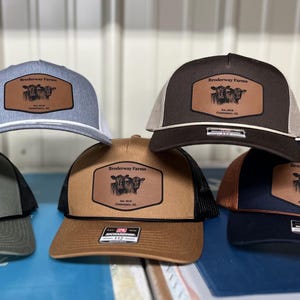 May include: A collection of six trucker hats in various colors, including gray, brown, and navy. Each hat features a leather patch with a cow illustration and the text "Broderway Farms" and "Est. 2016, Centreville, AL."